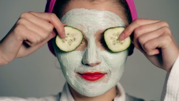 Close up van een meisje met een gezichtsmasker en een handdoek op haar hoofd die haar ogen met komkommers op een grijze achtergrond sluit. Face care concept. Mooi meisje met cosmetische masker op haar gezicht. — Stockvideo