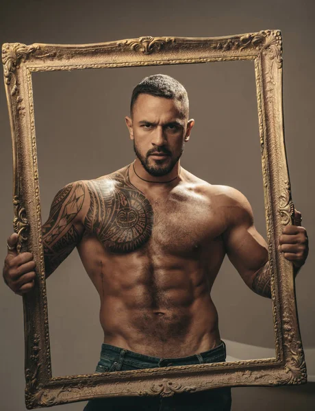 Latin Man with muscular body holds a frame and looks seriously. Muscular latin lover. Handsome brutal man on gray background. Portrait of brutal handsome male model.