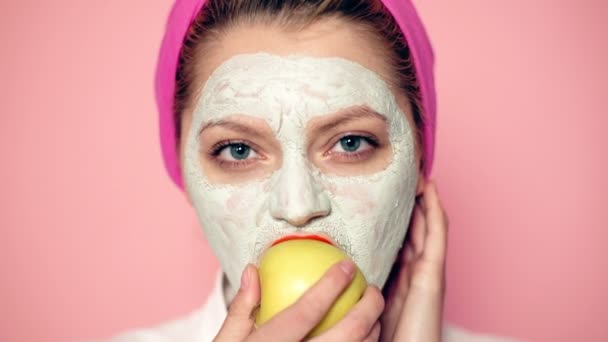 Close up van een meisje met een gezichtsmasker en een handdoek op haar hoofd die een appel op een roze achtergrond eet. Mooi meisje met cosmetische masker op haar gezicht. Vrouw dragen gezichtsmasker. Face care concept. — Stockvideo
