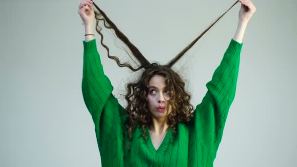 Curly girl with braces in a green sweater pulls her hair off on a white background. The concept of hair care. Parisian girl in winter clothes. Fashion look and beauty concept. — Stock Video