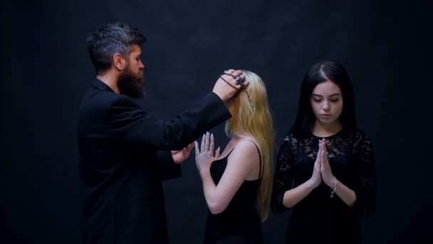 Prästen välsignar två tjejer på en svart bakgrund. Begreppet tillbedjan. Två flickor klädda i svart är erkände prästen. Mysterium bekännelse. Halloween party och fest koncept. — Stockvideo
