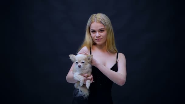 Blondynka z psem na rękach na czarnym tle. Dziewczyna z psem w studio. — Wideo stockowe