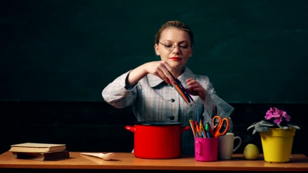 Ein Mädchen mit Gläsern am Tisch wirft Buntstifte und Stifte in einen Topf auf dem Hintergrund einer grünen Studentenbude. Konzept der Nahrung für das Gehirn. — Stockvideo