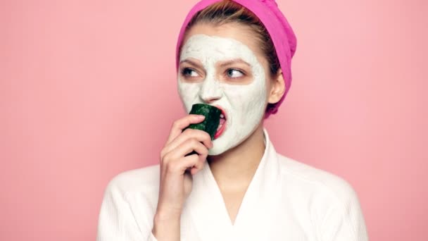 Uma menina com uma máscara no rosto e uma toalha na cabeça comendo um pepino em um fundo rosa. Menina bonita com máscara cosmética em seu rosto. Conceito de cuidado facial. — Vídeo de Stock