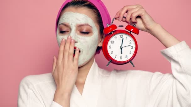 Sleeping girl with a mask on the face and a towel on his head holds the alarm clock in hes hand. Beautiful girl with cosmetic mask on her face. Woman wearing face mask. Face care concept. — Stock Video