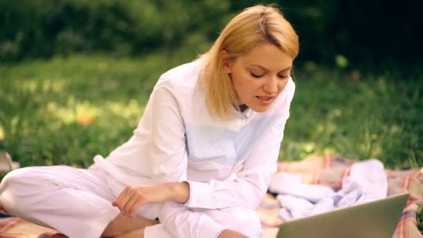 Girl on a lawn with a laptop. Undergraduate girl is making homework via the netbook while sitting on the meadow. — Stock Video