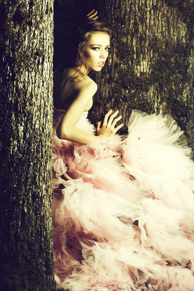 Beautiful young woman in pink glamour dress with long curly hair sitting on tree in sunny forest