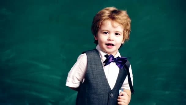 Un niño pequeño sostiene un bolígrafo en sus manos y luego lo esconde detrás de su espalda en el fondo de una pizarra de estudiantes verdes. Concepto de aprendizaje. Niños en uniforme. Profesor en el aula . — Vídeos de Stock