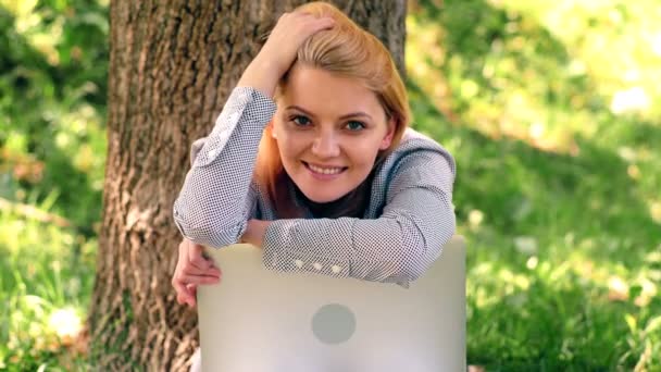 Een meisje op zoek naar de camera leunend op een laptop zittend op het gras in het park. Een student die rust in het park na studeren. — Stockvideo