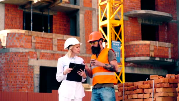 En kvinna i en vit hjälm och en skäggig man tala på byggplatsen. Begreppet konstruktion. Två ingenjörer diskuterar en byggnadsplan på byggarbetsplatsen. — Stockvideo