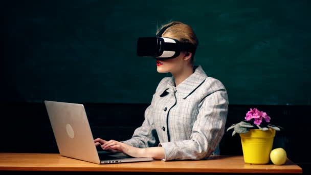 Eine Frau arbeitet an einem Laptop mit einer Virtual-Reality-Brille auf dem Hintergrund einer Studentenbude. das Konzept der virtuellen Realität. Lehrer mit Virtual-Reality-Brille am Tisch. — Stockvideo