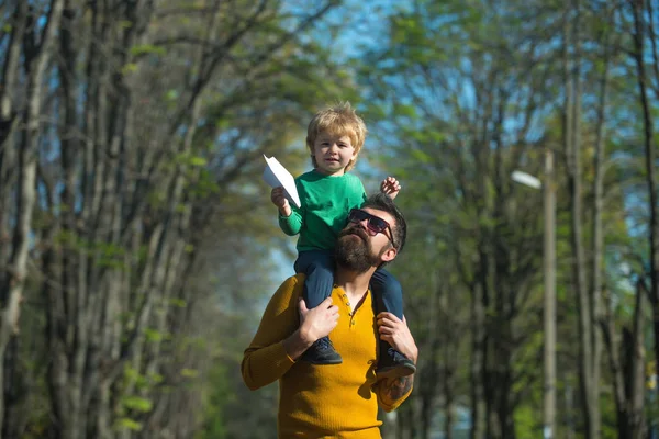 Voando como um pássaro. Pequeno filho sonha em voar no ombro dos pais. Menino e pai lançam avião de papel no parque . — Fotografia de Stock