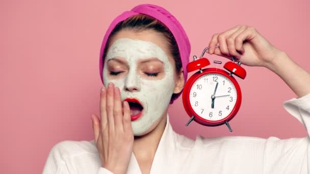 Een vrouw met een masker op haar gezicht en een handdoek op haar hoofd houdt een wekker in haar hand en gaapt op de roze achtergrond. Het concept van een gezond lichaam. — Stockvideo