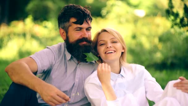那个留胡子的男人和女孩在公园的草地上坐着, 玩得很开心。白头发的女孩穿着白色的衣服坐在草地上, 有胡子的男子, 并在他的胡子上的叶子. — 图库视频影像