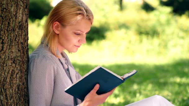 Het meisje leest van een notebook zit op het gras in het park. Meisje student bereidt voor klassen in het park. Het concept van buiten onderwijs. — Stockvideo