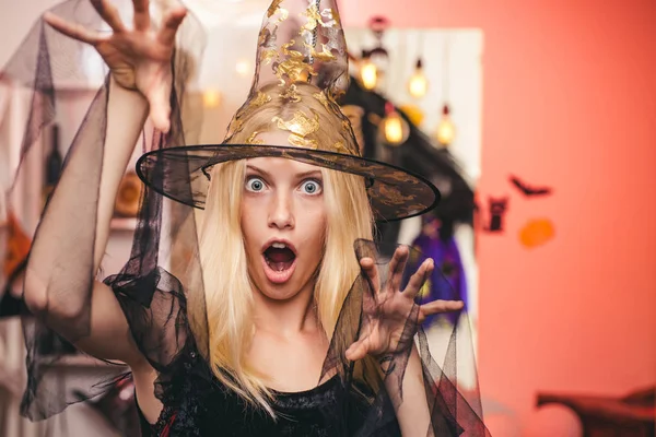 Belle jeune femme surprise en chapeau de sorcières et costume pointant la main montrant des produits. Belle jeune femme en costume de sorcière noire avec chapeau et balai debout sur fond orange. — Photo