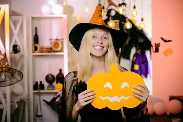 Griezelige kostuums van feestgangers. Mooie jonge blonde heks die op de bezem vliegt, geïsoleerd tegen een oranje achtergrond. glimlachende vrouw met mooi gezicht holding pompoen in studio op oranje achtergrond. — Stockfoto