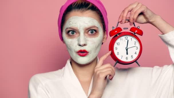 Kvinna med en mask på ansiktet och en handduk på huvudet håller en väckarklocka i händerna och leder ögonen i olika riktningar på rosa bakgrund. Face care koncept. — Stockvideo