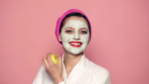 Meisje met een masker op haar gezicht en een handdoek op haar hoofd houdt een appel in haar handen en glimlacht naar de roze achtergrond. Begrip huidverzorging. — Stockvideo