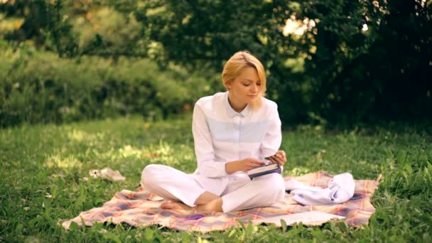 Chica vestida con ropa blanca hace notas en un cuaderno sentado en una manta en el parque. Estudiante estudiando en un parque . — Vídeo de stock