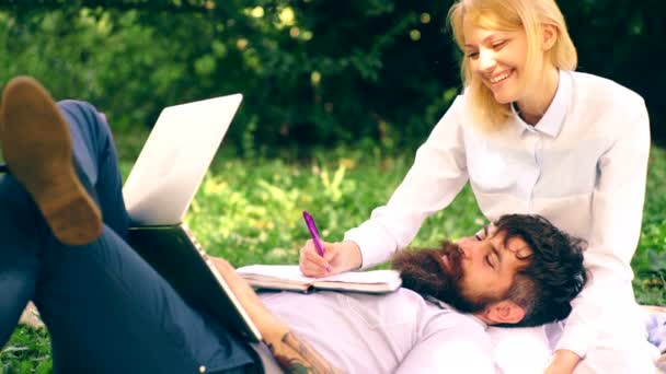 Pareja joven se prepara para los exámenes en el parque. Tipo barbudo y una chica bonita acostada en una manta con un portátil y anotar en un cuaderno en el parque. Concepto de entrenamiento en el parque . — Vídeo de stock