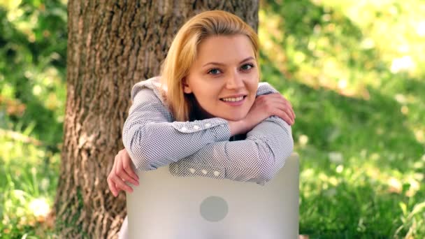 Дівчина сидить на траві в парку, спираючись на ноутбук і посміхається. мрійлива дівчина сидить в парку на траві . — стокове відео