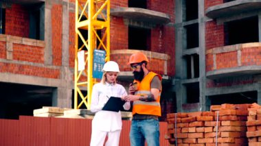Kasklı ve klasörlü bir kadın, inşaatın arka planına karşı koruyucu yelek giyen sakallı bir adamla iletişim kuruyor. Mühendis inşaat alanındaki müteahhitle iletişim kuruyor..