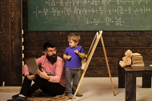 Profesor revisando el trabajo del estudiante en pizarra. El hombre que trabaja en el portátil habla con un niño pequeño. Concepto educativo — Foto de Stock