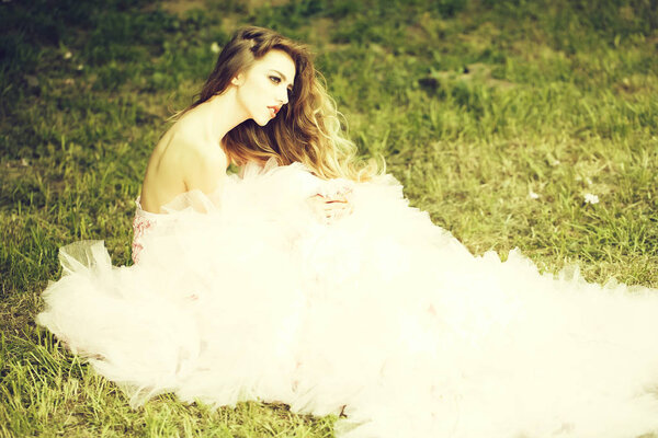 Beautiful young woman in pink glamour dress with long curly hair sitting on green grass in spring sunny day