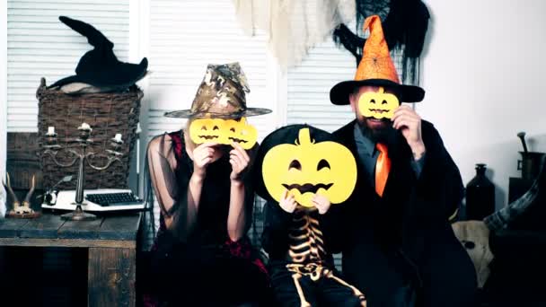 Женщина в костюме ведьмы, мужчина в костюме колдуньи и мальчик в костюме скелета прячутся за дыней на фоне декораций к Хэллоуину. Семья веселится на Хэллоуин . — стоковое видео