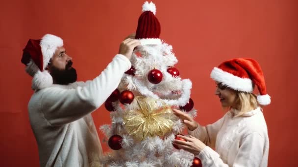 Pareja joven adorna el árbol de Navidad sobre fondo rojo. El hombre barbudo y la mujer hermosa en los sombreros de Año Nuevo adornan el árbol de Navidad sobre el fondo rojo. Concepto de Año Nuevo . — Vídeos de Stock