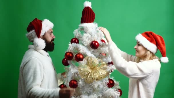 Pareja feliz decora un árbol de Navidad sobre un fondo verde. Barbudo hombre y mujeres rubias en Año Nuevo sombreros se divierten pasar tiempo decorando árbol de Navidad sobre fondo verde . — Vídeos de Stock