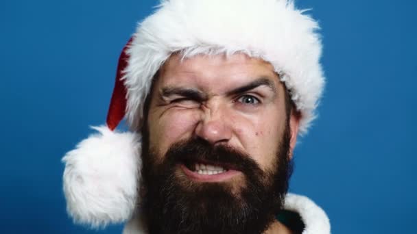 Homem barbudo no chapéu de Ano Novo faz caretas em um fundo azul. Pai Natal engraçado em um fundo azul. Conceito celebração ano novo . — Vídeo de Stock