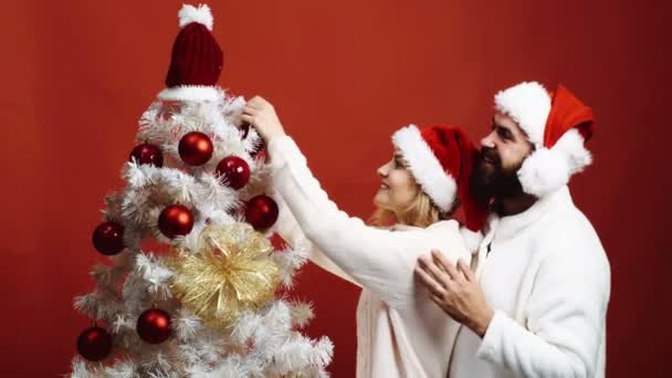 Una pareja joven adorna un árbol de Navidad sobre un fondo rojo. El hombre barbudo abraza a la mujer que decora el árbol de Navidad sobre fondo rojo. Concepto de Año Nuevo . — Vídeos de Stock