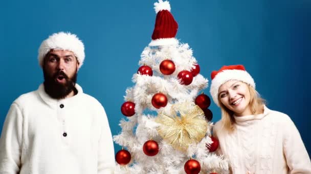 젊은 부부 grimaces 파란색 배경에 크리스마스 나무 근처에 서 게. 수염 난된 남자와 금발 머리 여자 나무 여 서 파란색 배경에 미소. 새 해 개념. — 비디오
