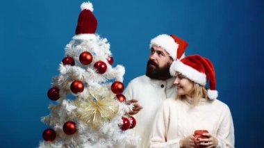 Sakallı adam ve yeni yıl şapka sarışın kadın mavi arka plan üzerinde Noel ağacı süsleyin. Sevgi dolu bir çift yeni yıl kutlama için hazırlanıyor. Yeni yıl kavramı.