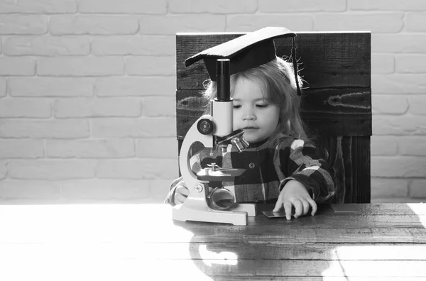 Criança inteligente em chapéu de graduação com estudo de microscópio no local de trabalho — Fotografia de Stock