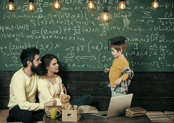 Parents listening their son, chalkboard on background. Homeschooling concept. Parents teaching kid at home. Boy presenting his knowledge to mom and dad. Smart child in graduate cap likes to study.