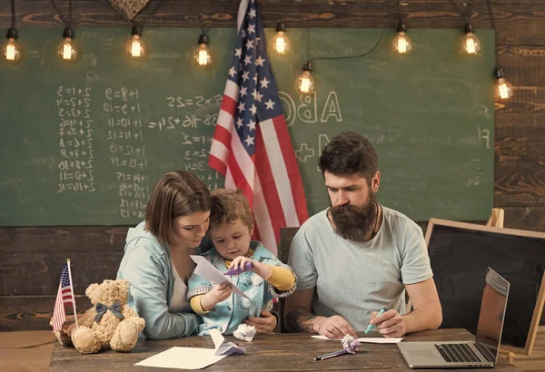American family at desk with son making paper planes. Homeschooling concept. Parents teaching son to create, handmade. Kid with parents in classroom with usa flag, chalkboard on background. — Stock Photo, Image