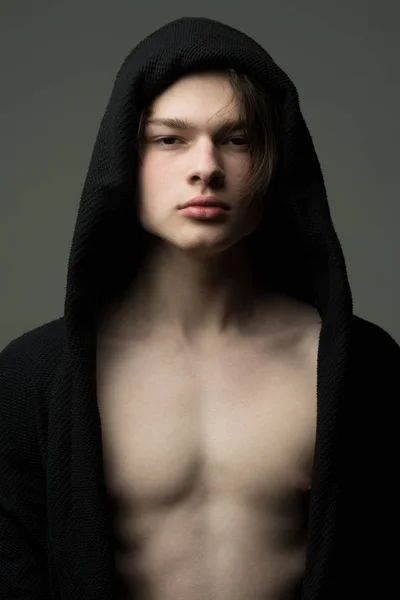 Tiener in zwarte badjas geïsoleerd op een grijze achtergrond. Closeup portret van gespierde jongeman met volle lippen en ernstige vertrouwen gezicht dragen van de motorkap, de mannelijkheid concept — Stockfoto