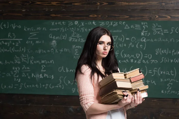 Serieuze jonge vrouwelijke student voor examens. Brunette meisje in glazen uitvoering twee stapels zware boeken — Stockfoto