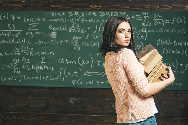 课程项目。女孩持有沉重的成堆的旧书, 黑板背景。女学生在科研方面工作。勤奋的学生准备考试测试。学生很喜欢学习 — 图库照片