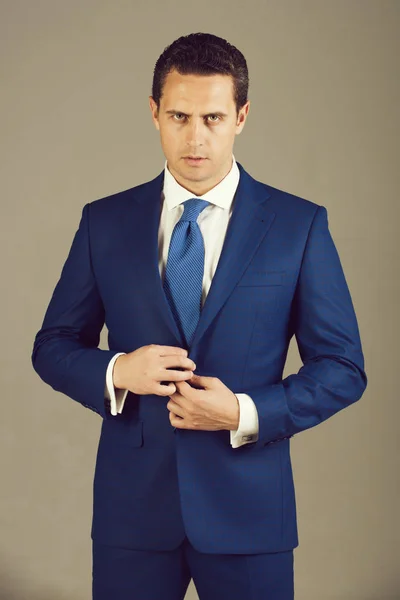 Handsome man, confident businessman or successful boss with stylish hair, haircut buttoning button on elegant blue formal suit jacket on grey background. Business, fashion and success. Dress code