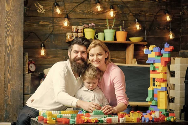 Kid with parents play with plastic blocks, build construction. Father, mother and cute son play with constructor bricks. Family leisure concept. Parents hugs, watching son playing, enjoy parenthood.