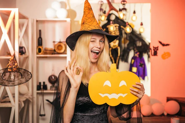 Beautiful young surprised woman in witches hat and costume holding pumpkin. Surprised woman in witches hat and short dress. Indoor portrait of cute young witch at halloween party.
