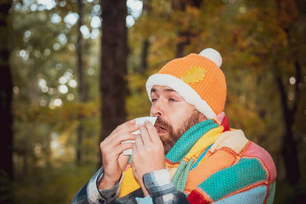 Mature man in jacket suffering from cold. Blowing nose with a tissue, looking miserable unwell very sick. Healthcare and medicine concept - ill man with paper tissue in autumn park. — Stock Photo, Image