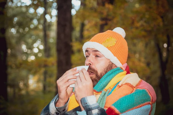 Hombre maduro con chaqueta que sufre de frío. Soplando la nariz con un pañuelo, pareciendo muy enfermo. Concepto de salud y medicina - hombre enfermo con tejido de papel en el parque otoñal . — Foto de Stock