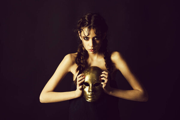 женщина с сияющим макияжем и косичками, держащая золотую маску
