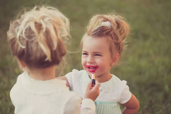 女の子は 緑の草の背景に妹のためのメイクアップをします 赤ちゃんは 子供モデルの唇に赤い口紅を適用します 美しさ ファッション スキンケア メイクアップの顔 幸せな子供 — ストック写真