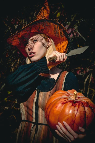 女性ゴシック魔女ハロウィーンの衣装と帽子。ハロウィーンの若い女性の肖像画の屋外の木製の背景に衣装します。カボチャをカットする準備をしての女の子. — ストック写真